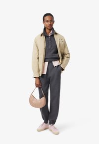 Veste isolante beige sur un pull gris, assortie à un pantalon de jogging gris foncé. Accessoirisée avec un sac à main matelassé rose et des baskets rose clair.