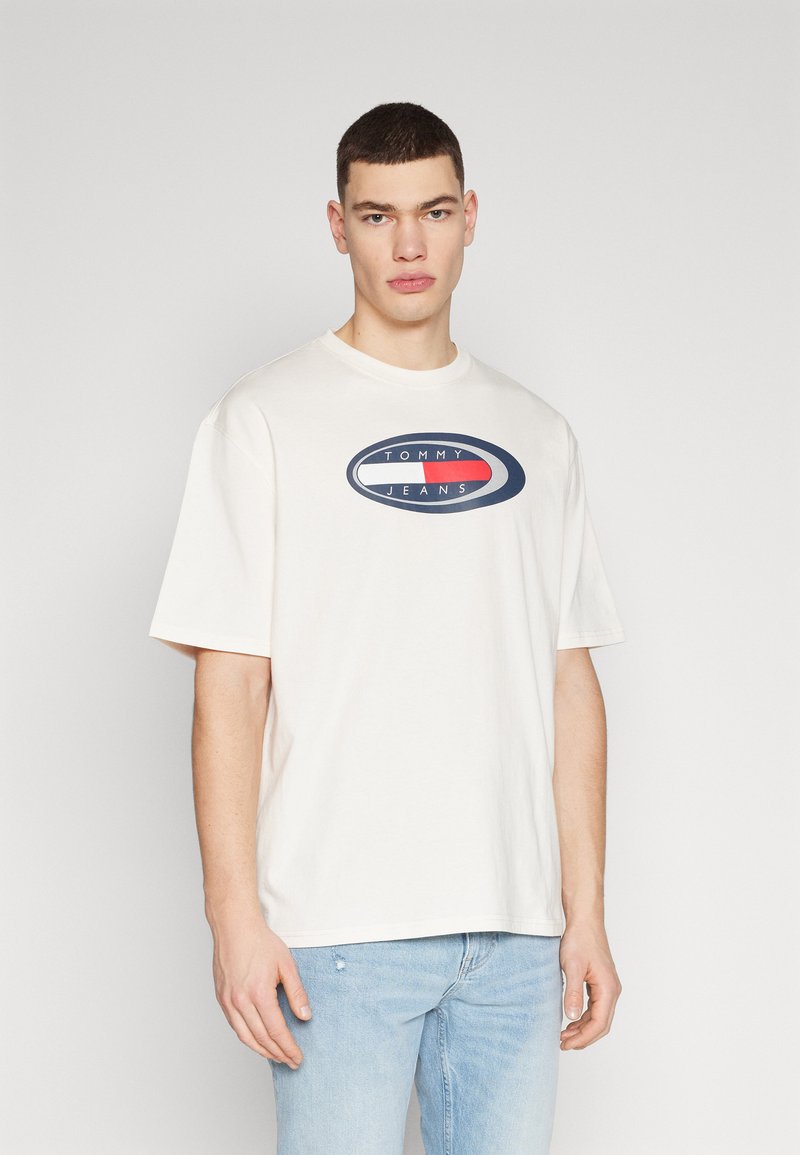 Camiseta de algodón blanca con cuello redondo y mangas cortas. Presenta un logo en el centro que dice "TOMMY JEANS" en azul marino, rojo y blanco.