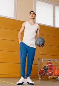 Athletic outfit featuring a sleeveless white top, blue pants, and white sneakers. The model holds a basketball in a gym setting.