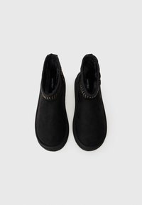 Botas de tornozelo pretas em camurça com um interior forrado macio, apresentando um painel texturizado com detalhes dourados à volta do tornozelo.