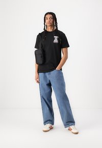 Svart bomulls t-skjorte med grafisk trykk, kombinert med løse blå denimjeans og hvite sneakers med røde striper. Liten svart crossover-bag.