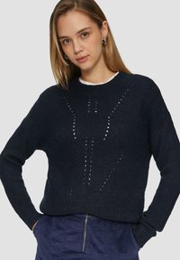 Marine gebreide trui met een ronde hals, textuurstof en opengewerkte details in een diagonaal patroon. Gedragen over een witte top.