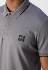 Polo gris avec un tissu texturé, col côte et détail boutonné. Présente un patch logo noir sur la poitrine gauche.