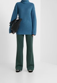 Pull à col roulé côtelé bleu, associé à un pantalon large texturé vert. Sac noir au design élégant et chaussures à bout pointu avec des accents.