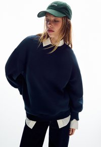 Námornická modrá oversized mikina s širokými rukávy, položená na bílé košili s límečkem, doplněná tmavozelenou baseballovou čepicí.