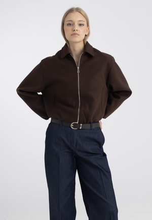 Jeune femme debout, les mains dans le dos, portant une veste zippée marron foncé et un pantalon bleu foncé avec une ceinture noire sur fond gris.