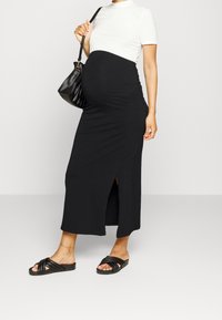 Svart maxikjol med en sidofick, kombinerad med en vit ribbad t-shirt med korta ärmar och svarta slide-sandaler. Handväska hängande över axeln.