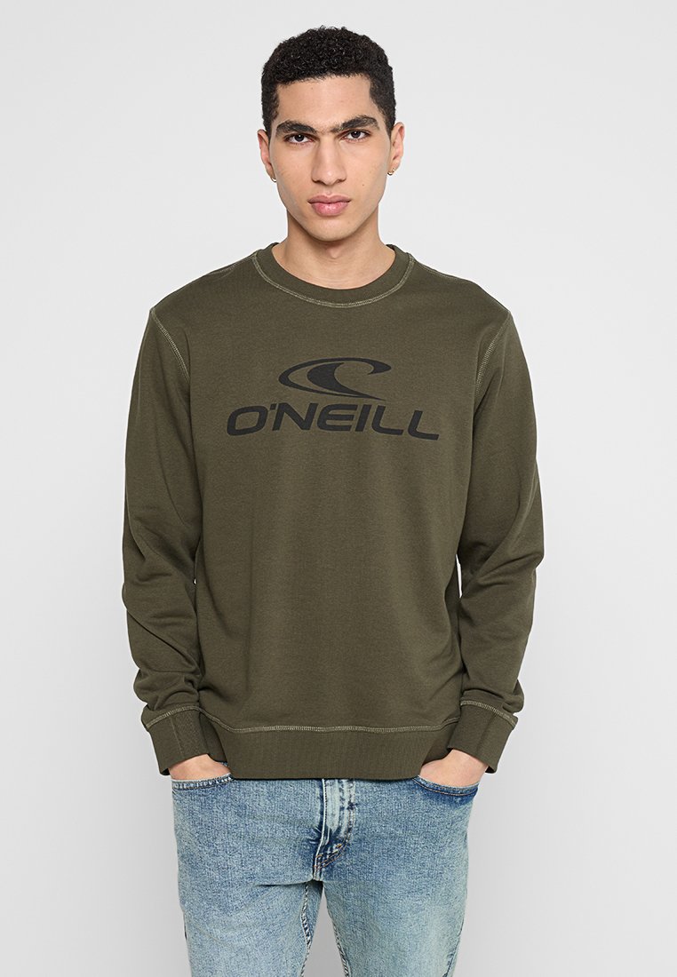Junger Mann, der einen olivgrünen O'Neill-Sweatshirt und hellblaue Jeans trägt, steht mit den Händen in den Taschen vor einem schlichten Hintergrund.