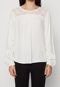 Vit blus med långa ärmar och detaljer av spets vid oket. Har samlade manschetter och en lös passform, tillverkad av ett mjukt och lätt tyg.