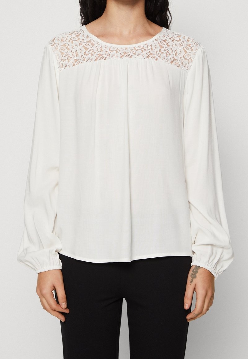 Vit blus med långa ärmar och detaljer av spets vid oket. Har samlade manschetter och en lös passform, tillverkad av ett mjukt och lätt tyg.