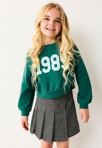 Usmievajúca sa dievčina s dlhými blond vlasmi, oblečená v zelenom mikinovom tričku s nápisom "1989" a šedom plisovanom sukni, stojaca na bielom pozadí.