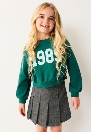 Ragazza sorridente con lunghi capelli biondi che indossa una felpa verde con la scritta "1989" e una gonna plissettata grigia, in piedi contro uno sfondo bianco.