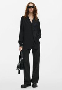 Camicia nera con bottoni e un'accenno di paillettes e dettagli di frange, abbinata a pantaloni neri a righe e scarpe nere chunky, con una piccola borsa nera.
