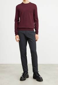 Homme portant un pull bordeaux ajusté, un pantalon slim gris foncé et des chaussures à semelle épaisse et lacets noires, debout sur un sol gris.