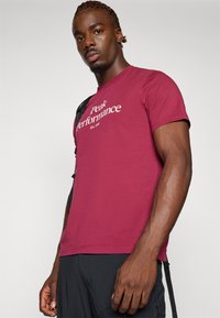 Bordeauxfarbene Baumwoll-T-Shirt mit kurzen Ärmeln, das auf der Vorderseite einen weißen Schriftzug "Peak Performance" und einen runden Ausschnitt hat.