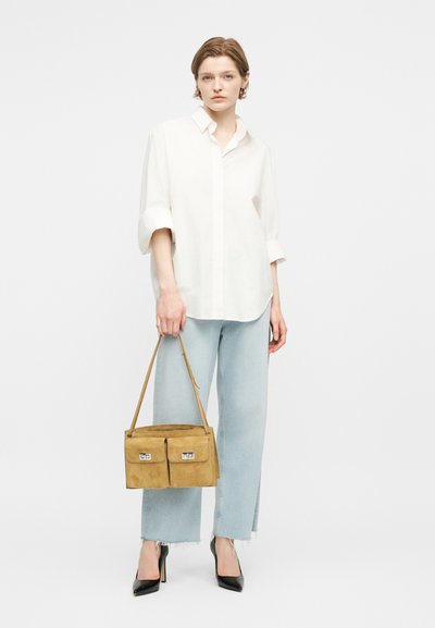 Γυναίκα που φοράει λευκό πουκάμισο και ανοιχτόχρωμα μπλε τζιν, κρατώντας μια καφέ τσάντα με δύο μπροστινές τσέπες, στέκεται μπροστά σε λευκό φόντο.