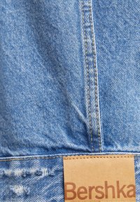 Ljust blå denimtyg med en strukturerad yta; har kontrasterande beige sömmar och en tanfärgad tagg som visar varumärket "Bershka."