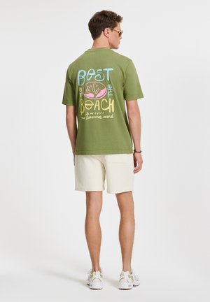 Olíva zöld t-shirt színes grafikus felirattal: "A LEGJOBB A TENGERNÉL" és pálma ikon a hátsó részen, krémszínű sorttal és sportcipővel kombinálva.