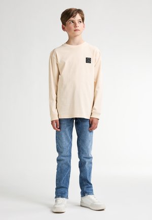 Chemise beige à manches longues avec un petit patch logo noir, associée à un jean bleu et des baskets blanches, se tenant devant un fond uni.