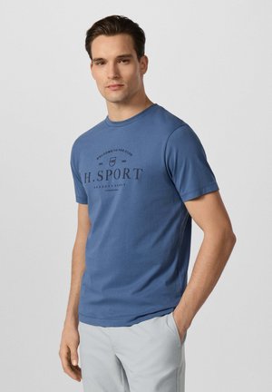 Junger Mann steht mit einer Hand in der Tasche, trägt ein blaues "H. Sport" T-Shirt und hellgraue Hose vor einfarbigem Hintergrund.