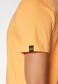 Orange bomulds T-shirt med korte ærmer, der har et sort mærke med gul logo på ærmet og ren syning. Glat tekstur.