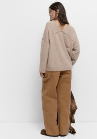 Beige strikketrøje med V-ryg, der har en løs pasform og ribbet kant, parret med løse brune bukser, som viser detaljer med sidelommer.