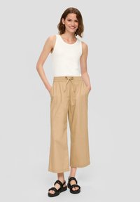 Beige wijde broek met een elastische koordtaille, gecombineerd met een witte tanktop en zwarte sandalen met dikke bandjes.