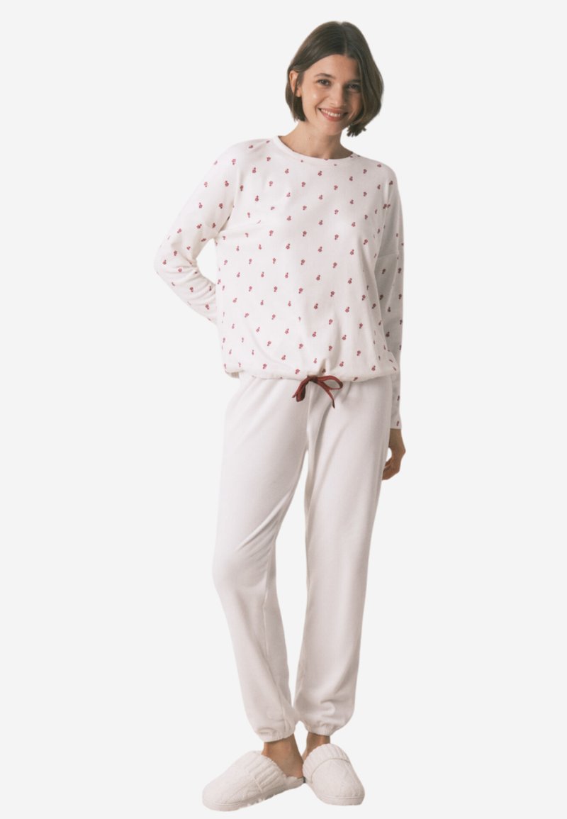 Ensemble de pyjama blanc comprenant un haut à manches longues avec des motifs floraux rouges et des poignets élastiques, associé à un pantalon fuselé et des chaussons pelucheux.