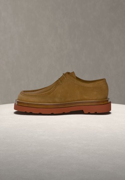 Καφέ καστόρινο καθημερινό παπούτσι με κορδόνια και παχιά κόκκινη λαστιχένια σόλα, εκτεθειμένο σε φωτεινή επιφάνεια με ουδέτερο φόντο.