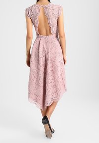 Vaaleanpunainen pitsimekko, jonka kuvio on saatu aikaan kaarrokeleikkauksesta, avonaisesta selkäosasta ja kerroksellisesta muotoilusta. Mekossa on levenevä helma ja monimutkaisia kuvioita.