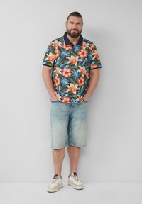 Heren polo met korte mouwen in marineblauw en een kleurrijke bloemenprint, gecombineerd met lichtblauwe denimshorts. Schoenen zijn wit met accenten.