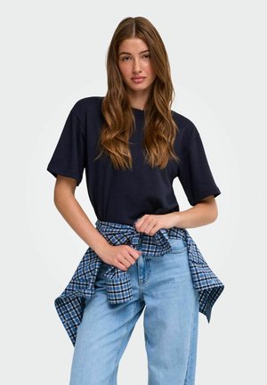 Jeune femme aux longs cheveux ondulés porte un t-shirt noir et un jean bleu clair avec une chemise à carreaux bleue nouée autour de sa taille sur un fond uni.