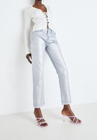 Jeans rectos metálicos plateados con cintura alta, combinados con una blusa blanca de manga larga con un frente con cordones, y tacones rosas con tiras.