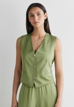 Kobieta o długich ciemnych włosach, ubrana w bezrękawnik i spodnie w kolorze oliwkowej zieleni z guzikami, na tle jasnego, jednolitego tła.