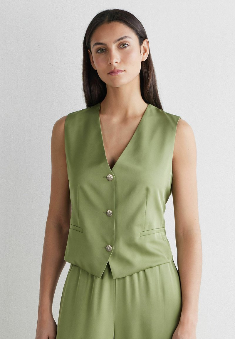 Vrouw met lang donker haar, gekleed in een mouwloos olijfgroen vest met knopen en bijpassende broek, tegen een effen lichte achtergrond.