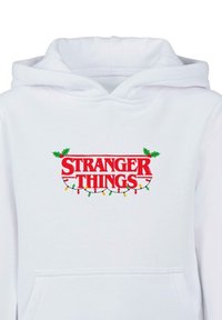 Bílá mikina s kapucí s nápisem "STRANGER THINGS" červeným textem a zelenými akcenty, zdobená cesmínou a barevnými vánočními světélky. Obsahuje přední kapsu.