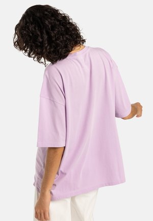 Person with curly dark hair wearing a loose light purple short-sleeve shirt and white pants, shown from the back and slightly turned.