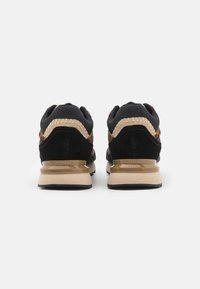 Zapatillas de ante negras y marrones con un diseño texturizado, que cuentan con una suela beige y costuras decorativas. La vista trasera muestra un detalle de la marca.