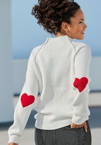 Witte geribbelde trui met rode hartpatches op de ellebogen, met een hoge nek en een aansluitend ontwerp. G gedragen met donkere jeans.