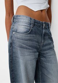 Närbild av midjan som bär blekta högmidjade blå jeans och en vit kort topp som visar bar hud ovanför midjebandet.