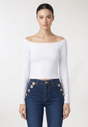 Młoda kobieta ubrana w biały, odkrywający ramiona, długi rękaw oraz ciemnoniebieskie, wysokowyste dżinsy z ozdobnymi złotymi guzikami.