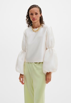 Žena s kučeravými vlasmi v mokrom vzhľade, oblečená v bielej blúzke s nafúknutými rukávmi, svetlozelených nohaviciach a hrubom zlatom reťazovom náhrdelníku.