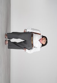 Moteris stovi prieš paprastą foną, vilkėdama baltą švarką, pilkas platėjančias kelnes su 'Moschino' diržu, rožinę skarelę ir juodus sandalus.