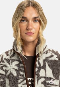 Veste en polaire crème et marron avec un motif floral, présentant une bordure marron, une fermeture à boutons et un patch logo sur le côté gauche.