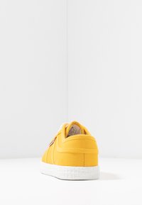 Sneaker in tela gialla con suola in gomma bianca, punta rotonda e cuciture a contrasto. Presenta una patche con logo sul lato.