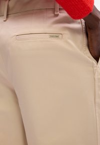 Bež hlače glatke teksture, s stražnjim džepom i malom oznakom Calvin Klein. Uočljivo šivanje uz šavove.