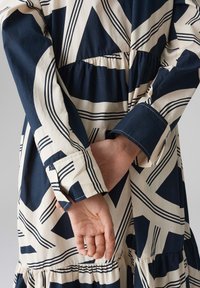 Blue and cream dress featuring geometric patterns, gathered waist, and long sleeves with buttoned cuffs. Smooth cotton fabric.