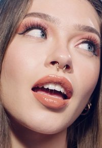 Jeune femme aux yeux bleu clair, rouge à lèvres pêche brillant, longs cils et anneau de septum en or, regardant sur le côté avec la bouche légèrement ouverte.