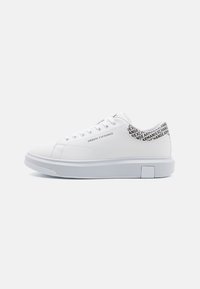 Zapatillas blancas con parte superior de cuero sintético suave, punta redonda y suela de goma blanca plana. La parte trasera presenta un patrón de texto negro con "Armani Exchange."