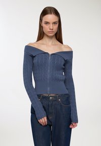 Niebieski sweter bez ramiączek z zamkiem, z fakturą w warkocz, ściągaczami na mankietach oraz dopasowanym krojem, w zestawie z ciemnoniebieskimi dżinsami.